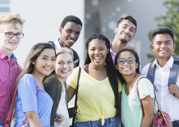A group of eight multi-ethnic teenagers, 17 and 18 years old, carrying book bags, standing together outside a school building. They are high school seniors or university freshmen.