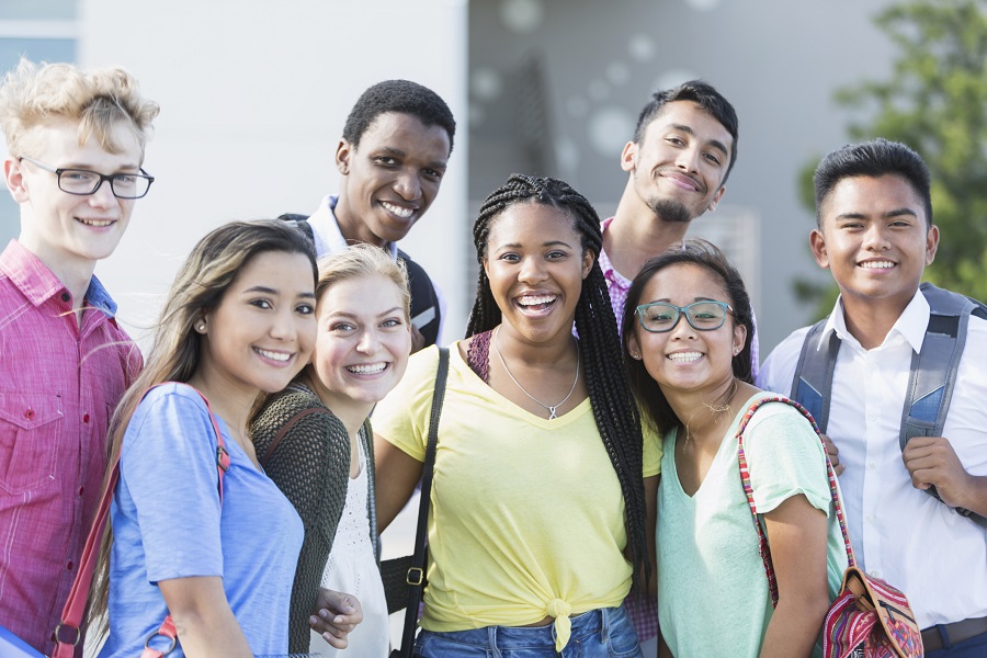 A group of eight multi-ethnic teenagers, 17 and 18 years old, carrying book bags, standing together outside a school building. They are high school seniors or university freshmen.